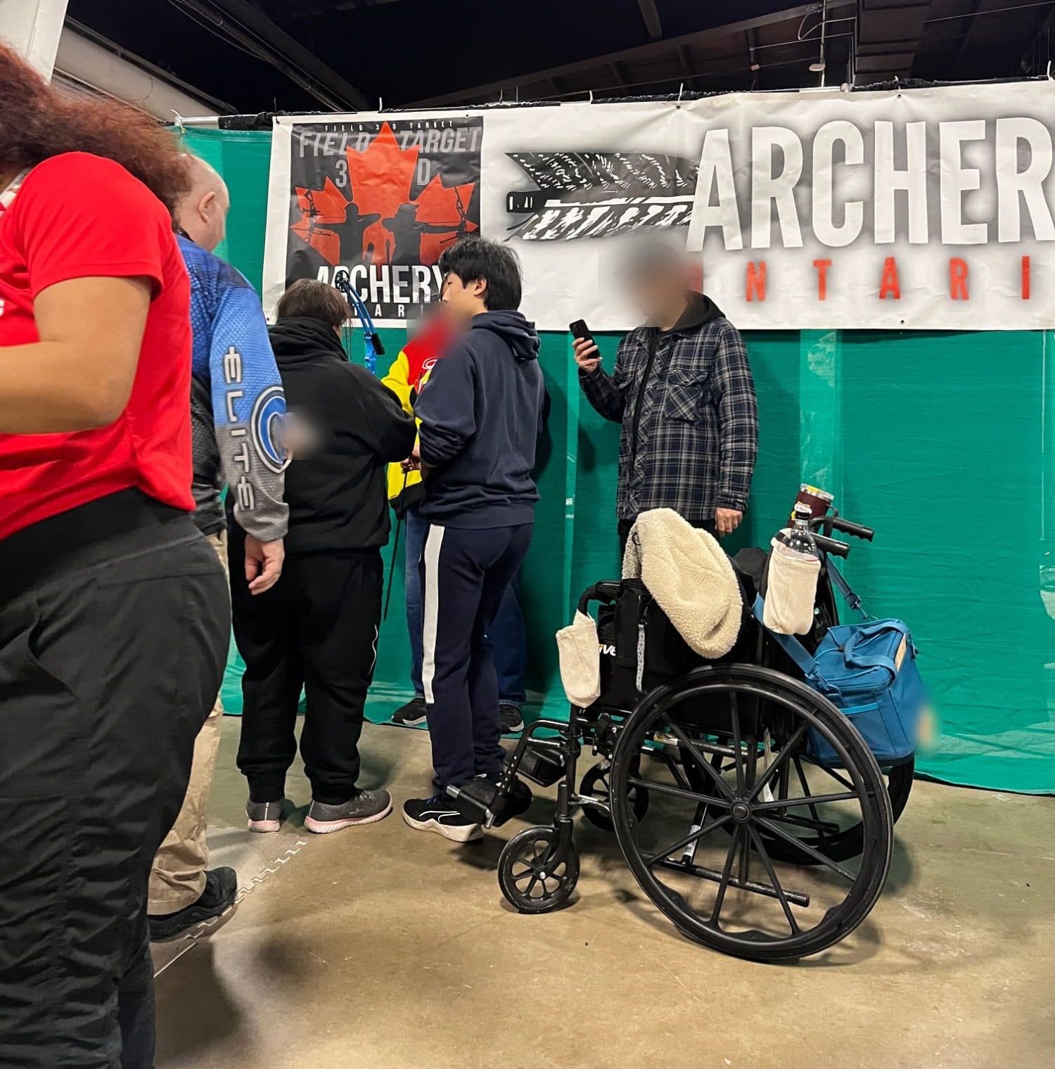 The Archery Ontario booth at a show, with a founder helping an archer. Other faces are blurred.
