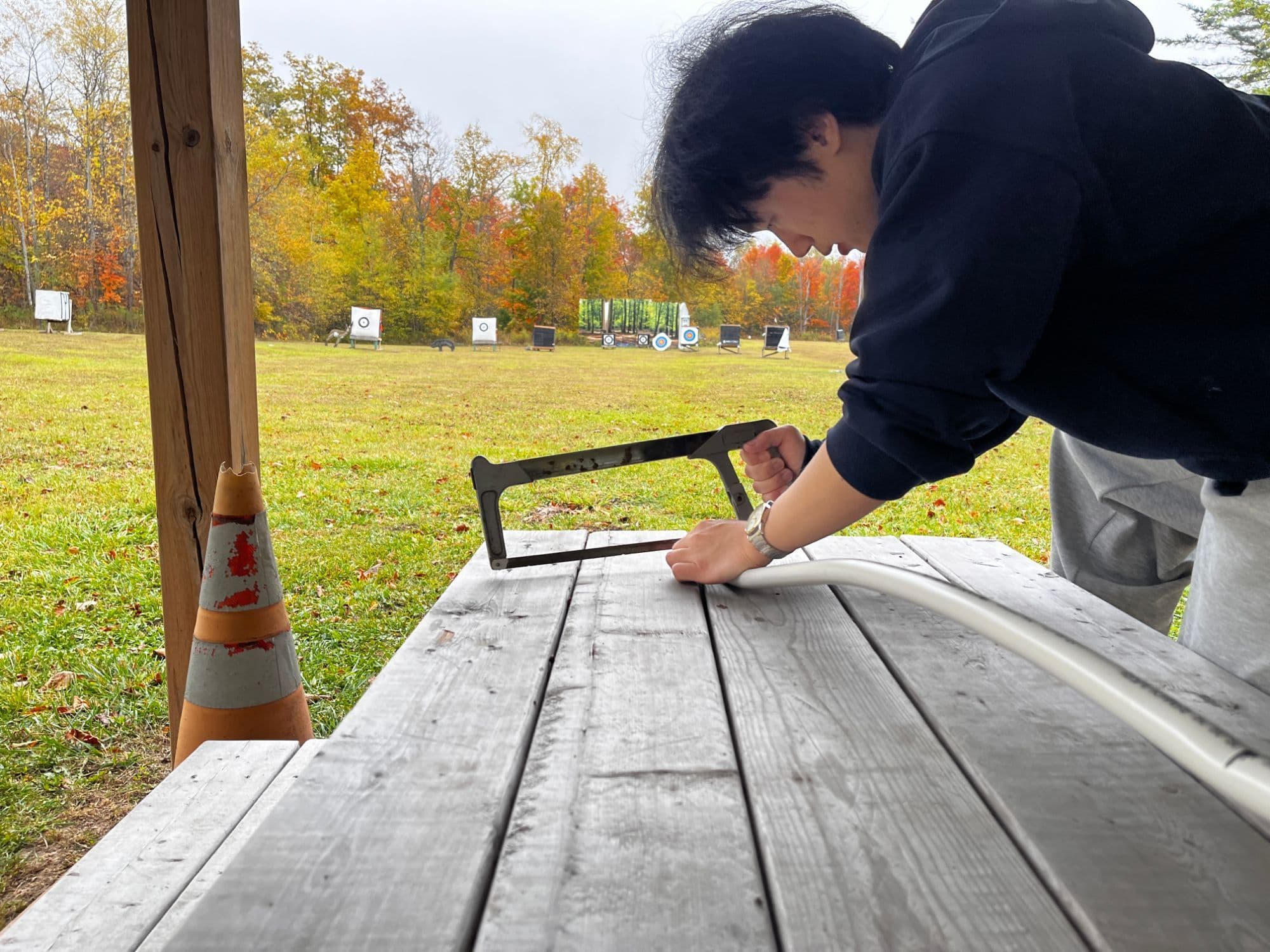 A founder cutting white PVC pipe with a hacksaw on a picnic table at an outdoor archery range, autumn trees and targets behind.