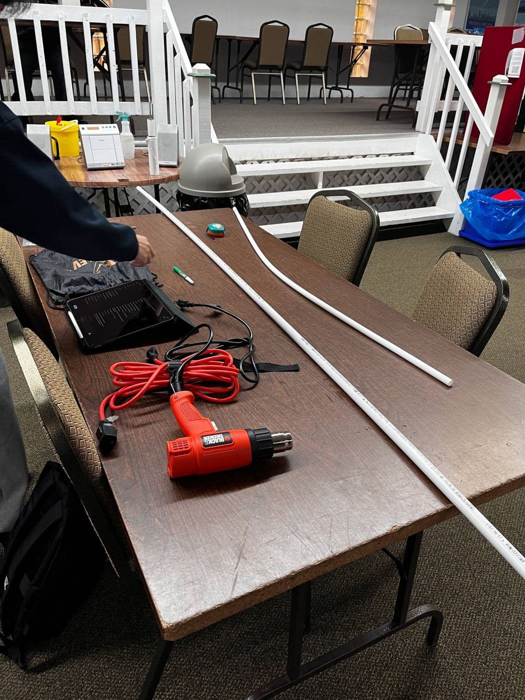 Two lengths of white PVC pipe on a table, one already bent, next to an orange heat gun, a tape measure and a marker.