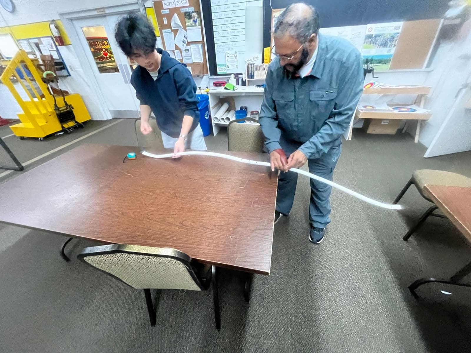 Two people holding a bent PVC bow across a table to check its curve.
