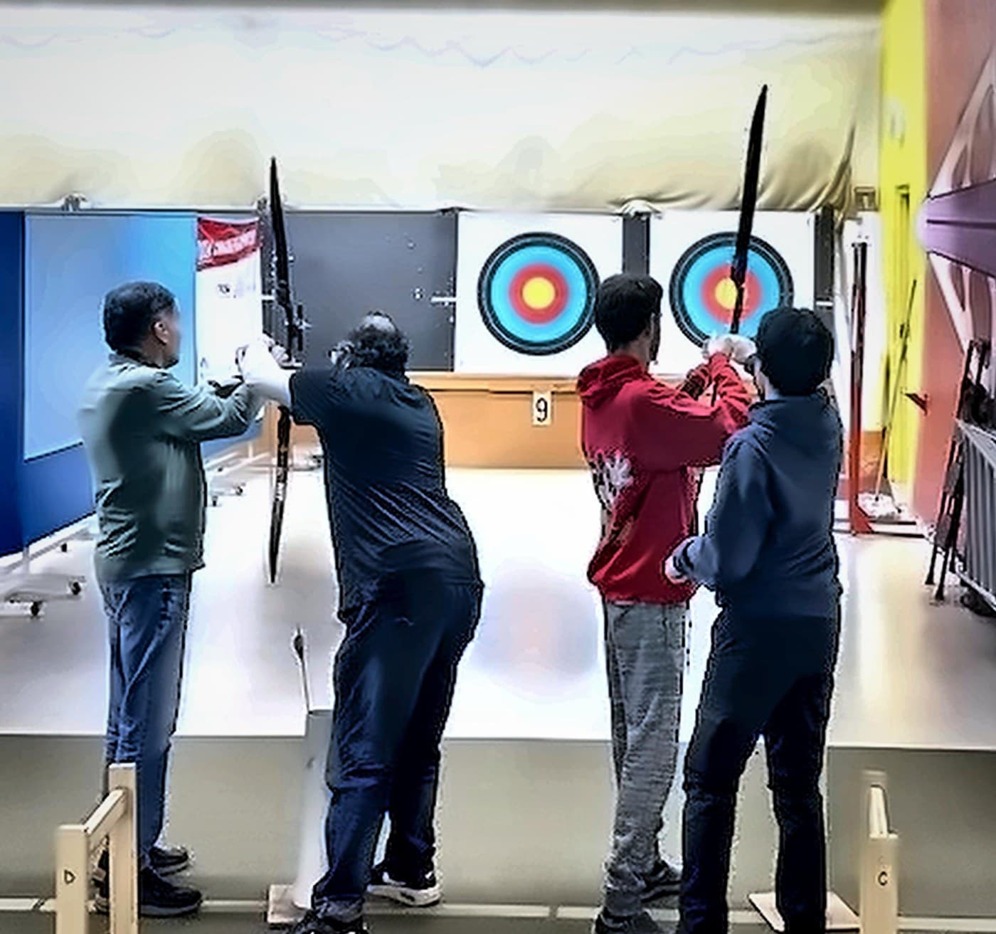 A wide view from behind of an adaptive archery session at the range: four archers on the line drawing recurve bows toward two targets.